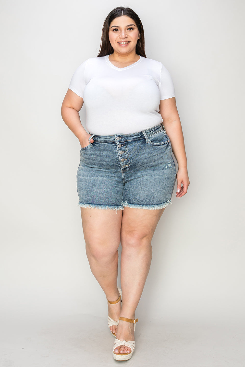 Wearing the Judy Blue Full Size Button Fly Raw Hem Denim Shorts and a white t-shirt, a person stands smiling in sandals against a plain background.