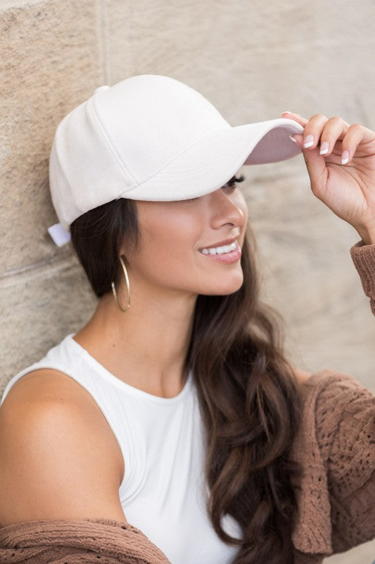 A smiling woman wearing a structured crown Velour Ball Cap and a brown cardigan leans against a stone wall.