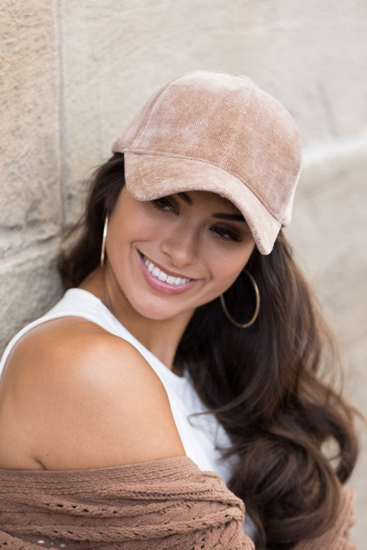 A smiling woman wearing a structured crown Velour Ball Cap and a brown cardigan leans against a stone wall.