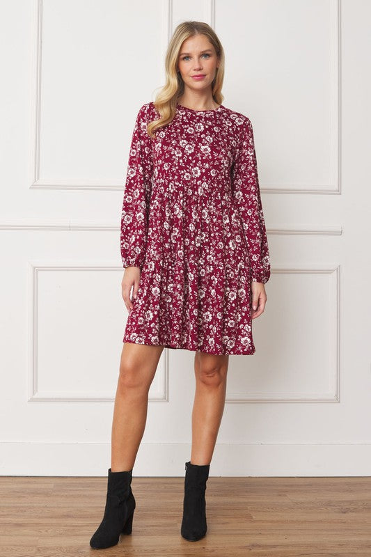 A woman in a Floral Crew Neck Midi Dress stands indoors against a white paneled wall. She is smiling, with one hand by her side and the other holding part of her dress. The red floral dress, made of polyester with a hint of spandex, complements her black ankle boots perfectly.