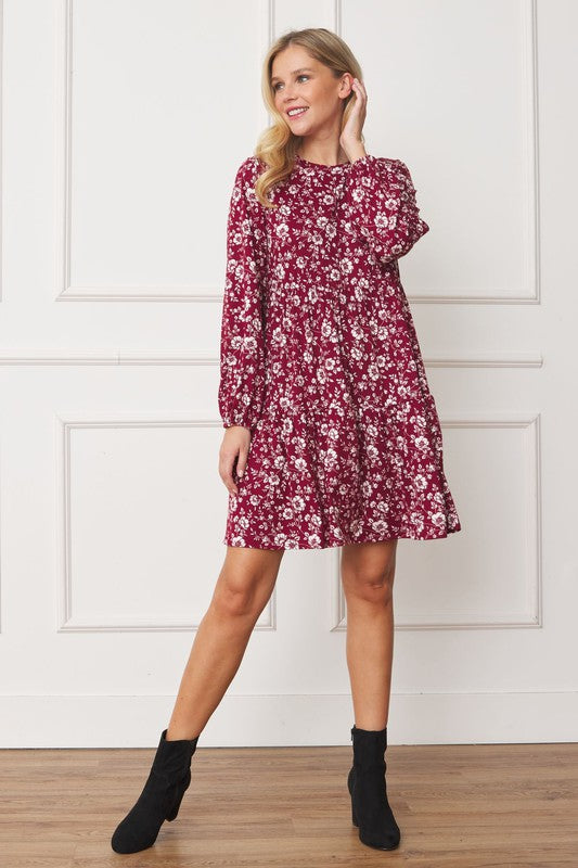 A woman in a Floral Crew Neck Midi Dress stands indoors against a white paneled wall. She is smiling, with one hand by her side and the other holding part of her dress. The red floral dress, made of polyester with a hint of spandex, complements her black ankle boots perfectly.