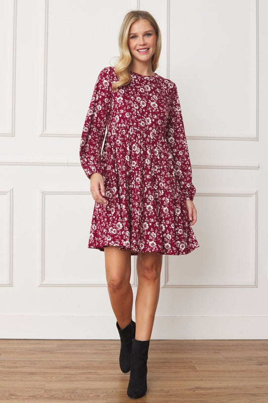 A woman in a Floral Crew Neck Midi Dress stands indoors against a white paneled wall. She is smiling, with one hand by her side and the other holding part of her dress. The red floral dress, made of polyester with a hint of spandex, complements her black ankle boots perfectly.