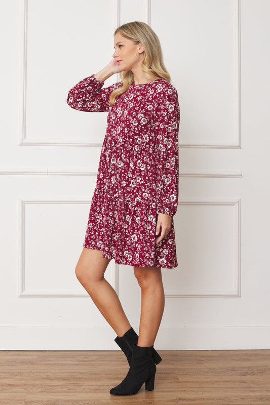 A woman in a Floral Crew Neck Midi Dress stands indoors against a white paneled wall. She is smiling, with one hand by her side and the other holding part of her dress. The red floral dress, made of polyester with a hint of spandex, complements her black ankle boots perfectly.