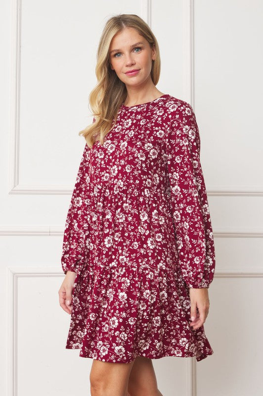 A woman in a Floral Crew Neck Midi Dress stands indoors against a white paneled wall. She is smiling, with one hand by her side and the other holding part of her dress. The red floral dress, made of polyester with a hint of spandex, complements her black ankle boots perfectly.