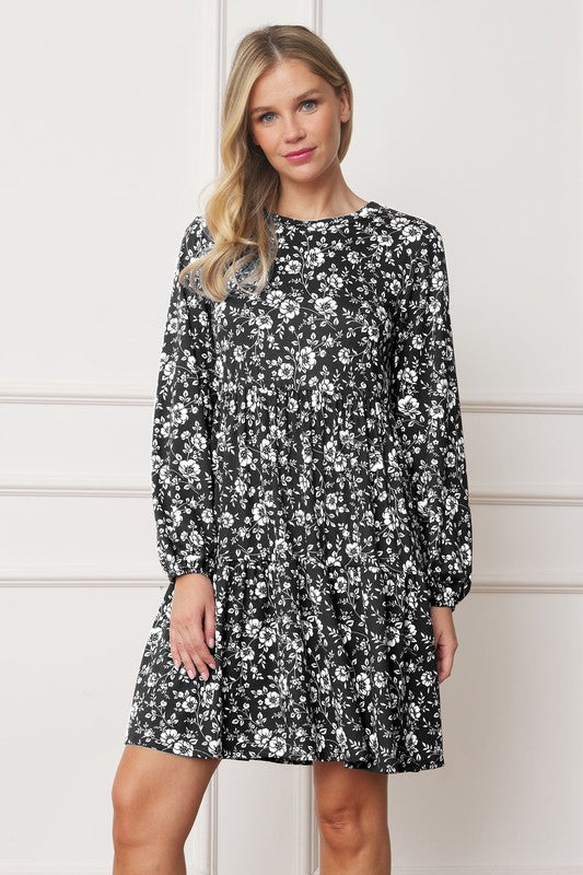 A woman in a Floral Crew Neck Midi Dress stands indoors against a white paneled wall. She is smiling, with one hand by her side and the other holding part of her dress. The red floral dress, made of polyester with a hint of spandex, complements her black ankle boots perfectly.