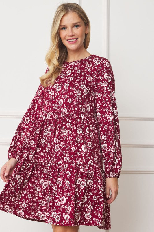 A woman in a Floral Crew Neck Midi Dress stands indoors against a white paneled wall. She is smiling, with one hand by her side and the other holding part of her dress. The red floral dress, made of polyester with a hint of spandex, complements her black ankle boots perfectly.