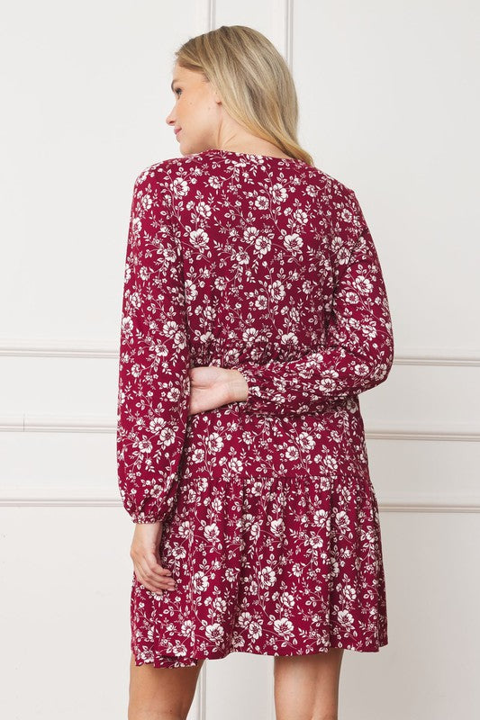 A woman in a Floral Crew Neck Midi Dress stands indoors against a white paneled wall. She is smiling, with one hand by her side and the other holding part of her dress. The red floral dress, made of polyester with a hint of spandex, complements her black ankle boots perfectly.