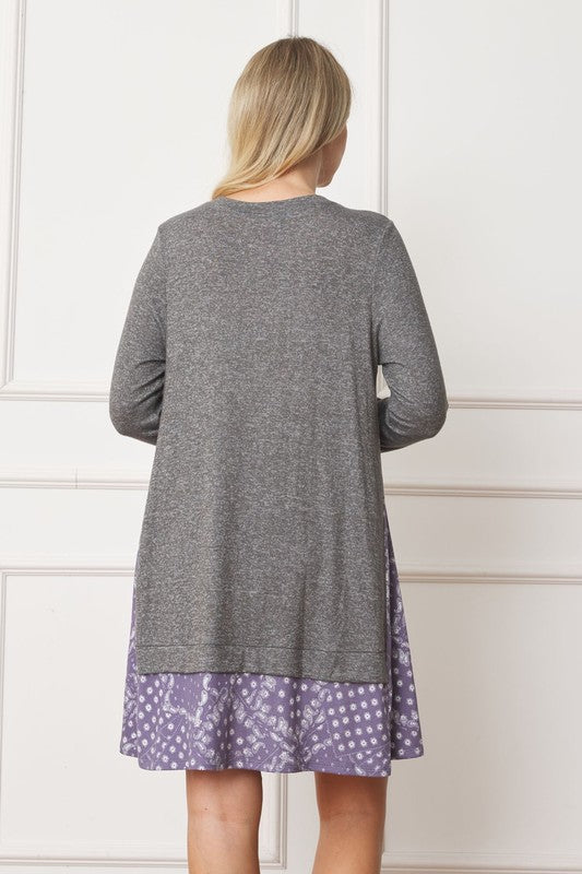 A woman with blonde hair is wearing a Mock Layered Bandana Paisley Mini Dress, featuring long sleeves and patterned fabric at the hem. She is standing indoors against a white wall.