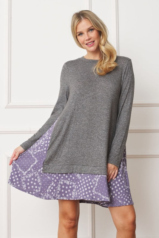 A woman with long blonde hair wears a grey long-sleeve sweater layered over the Mock Layered Bandana Paisley Mini Dress. She is standing indoors against a plain white wall and looking downward.