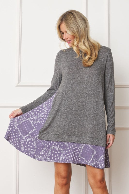 A woman with long blonde hair wears a grey long-sleeve sweater layered over the Mock Layered Bandana Paisley Mini Dress. She is standing indoors against a plain white wall and looking downward.