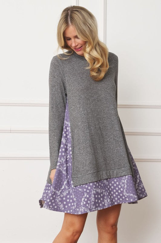 A woman with long blonde hair wears a grey long-sleeve sweater layered over the Mock Layered Bandana Paisley Mini Dress. She is standing indoors against a plain white wall and looking downward.