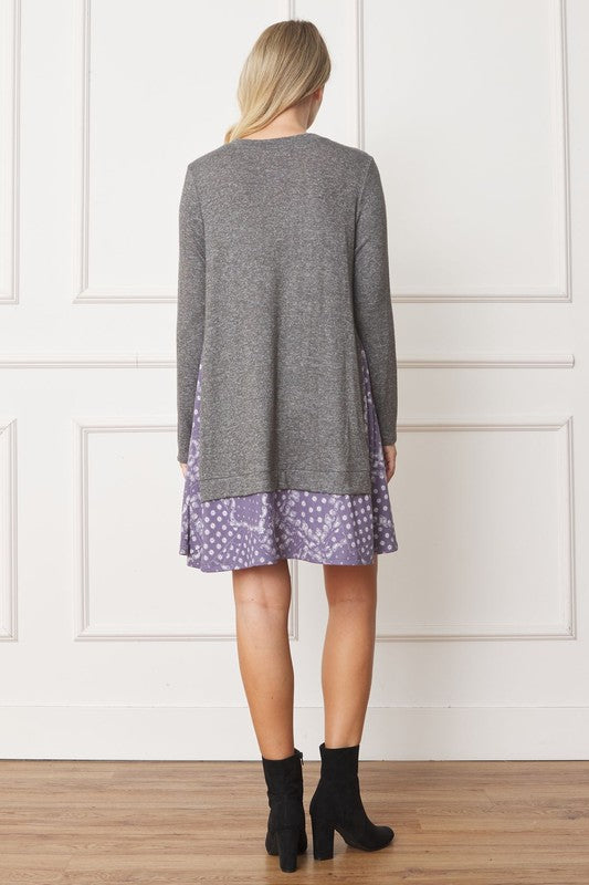 A woman with long blonde hair wears a grey long-sleeve sweater layered over the Mock Layered Bandana Paisley Mini Dress. She is standing indoors against a plain white wall and looking downward.