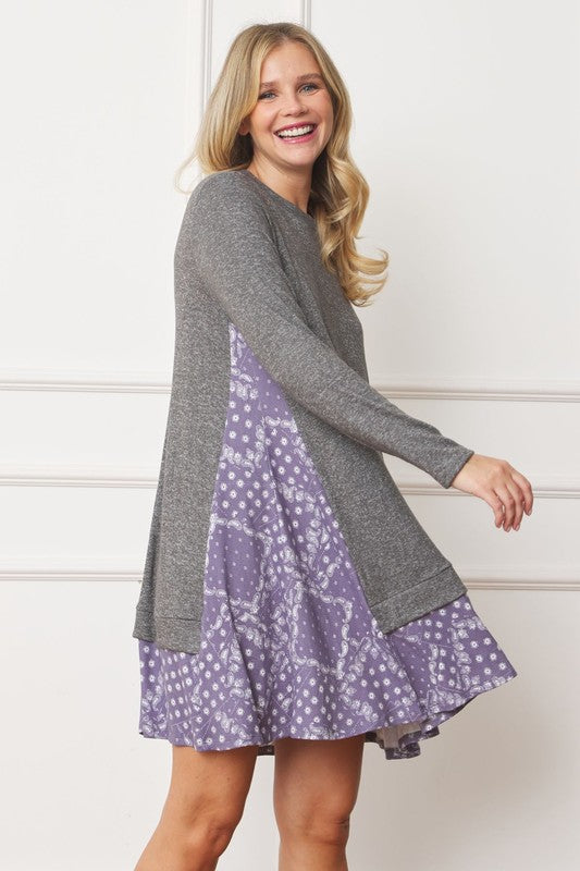 A woman with blonde hair is wearing a Mock Layered Bandana Paisley Mini Dress, featuring long sleeves and patterned fabric at the hem. She is standing indoors against a white wall.