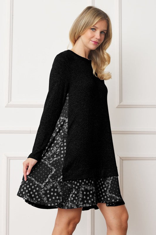 A woman with blonde hair is wearing a Mock Layered Bandana Paisley Mini Dress, featuring long sleeves and patterned fabric at the hem. She is standing indoors against a white wall.