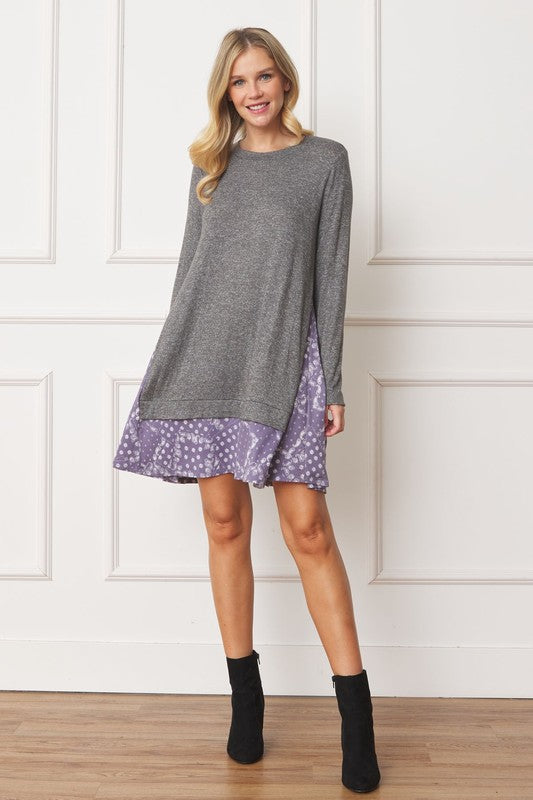 A woman with long blonde hair wears a grey long-sleeve sweater layered over the Mock Layered Bandana Paisley Mini Dress. She is standing indoors against a plain white wall and looking downward.