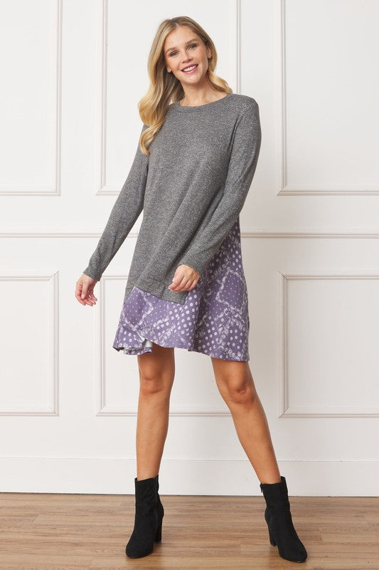 A woman with blonde hair is wearing a Mock Layered Bandana Paisley Mini Dress, featuring long sleeves and patterned fabric at the hem. She is standing indoors against a white wall.