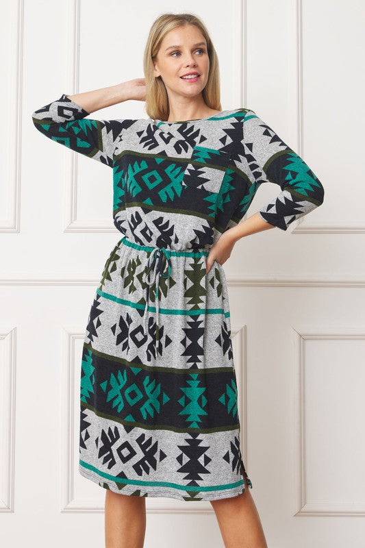 A woman in a green, black, and gray Plus Tribal Drawstring Blouson Midi Dress stands with one hand on her hip and the other behind her head, posing in front of a white paneled wall.