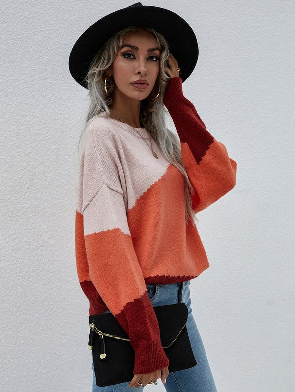 A woman with long, wavy hair wears the Women's Long Sleeve Round Neck Sweater in color-blocked shades of pink, orange, and red. She pairs it with jeans and a black hat, holds a black clutch, and stands against a plain, light-colored background.