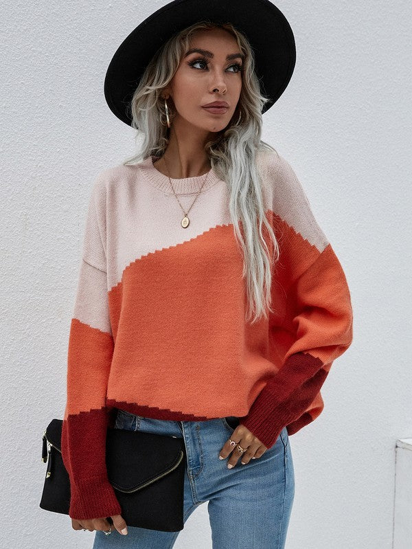 A woman with long, wavy hair wears the Women's Long Sleeve Round Neck Sweater in color-blocked shades of pink, orange, and red. She pairs it with jeans and a black hat, holds a black clutch, and stands against a plain, light-colored background.