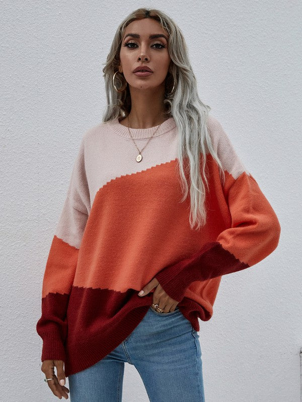 A woman with long, wavy hair wears the Women's Long Sleeve Round Neck Sweater in color-blocked shades of pink, orange, and red. She pairs it with jeans and a black hat, holds a black clutch, and stands against a plain, light-colored background.