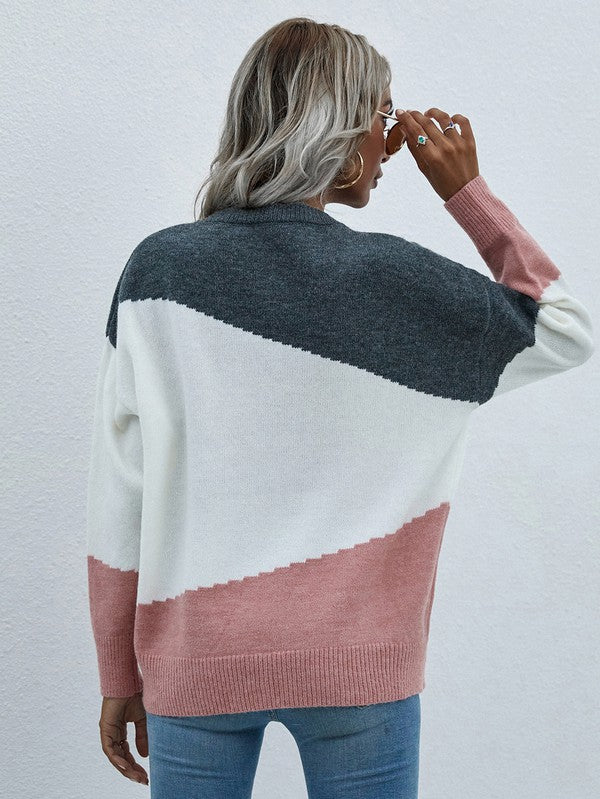 A woman with long, wavy hair wears the Women's Long Sleeve Round Neck Sweater in color-blocked shades of pink, orange, and red. She pairs it with jeans and a black hat, holds a black clutch, and stands against a plain, light-colored background.