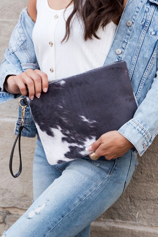 A person holding a Faux Fur Cow Animal Print Clutch with a leather strap, wearing a black top, light blue jeans, and a gold necklace.