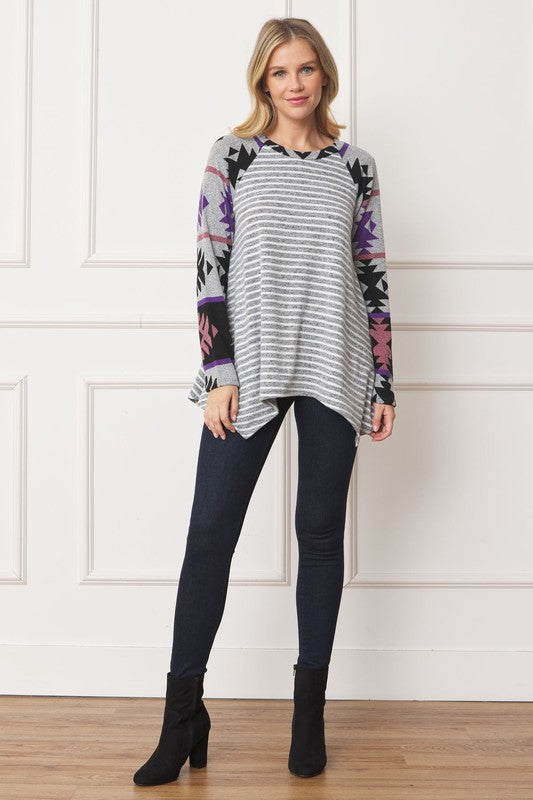 A woman with long blond hair, adorned in a gray, striped Tribal Sleeve Handkerchief Tunic featuring tribal sleeve patterns and dark jeans, is standing indoors in front of a paneled wall.