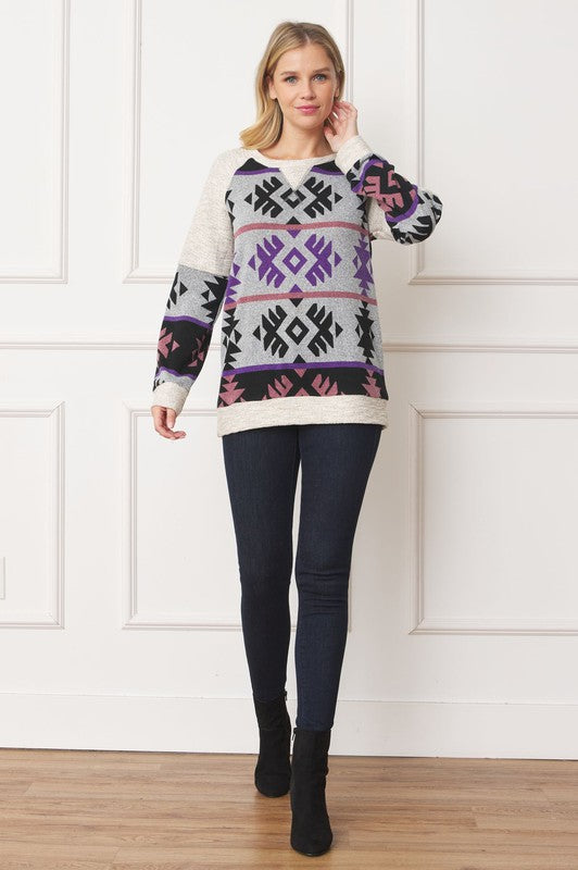 A woman stands smiling, wearing a Plus V Stitch Tribal Contrast Tunic with geometric patterns in black, grey, purple, and pink. The long-sleeve knit fabric is infused with gold thread. She has blonde hair and dark pants against a light-colored wall.