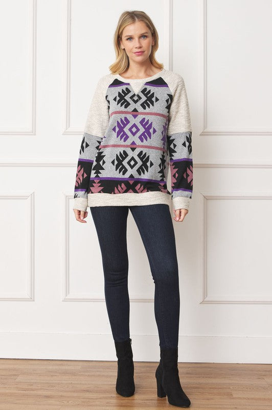 A woman stands smiling, wearing a Plus V Stitch Tribal Contrast Tunic with geometric patterns in black, grey, purple, and pink. The long-sleeve knit fabric is infused with gold thread. She has blonde hair and dark pants against a light-colored wall.