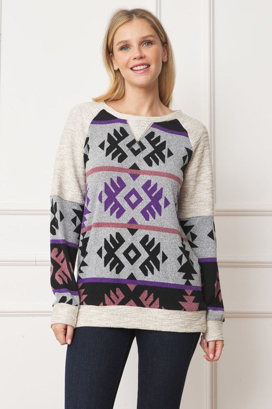 A smiling blonde-haired woman is wearing the V Stitch Tribal Contrast Tunic, which features geometric designs in purple, black, and pink, standing against a white background.