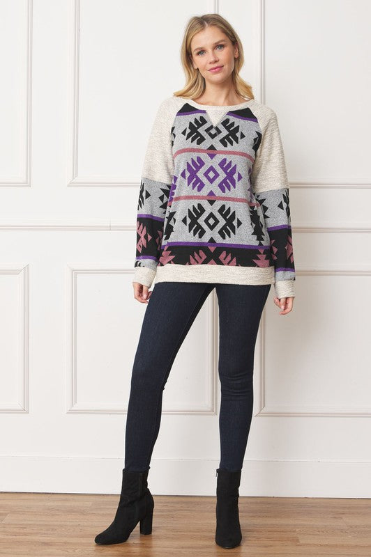A woman stands smiling, wearing a Plus V Stitch Tribal Contrast Tunic with geometric patterns in black, grey, purple, and pink. The long-sleeve knit fabric is infused with gold thread. She has blonde hair and dark pants against a light-colored wall.