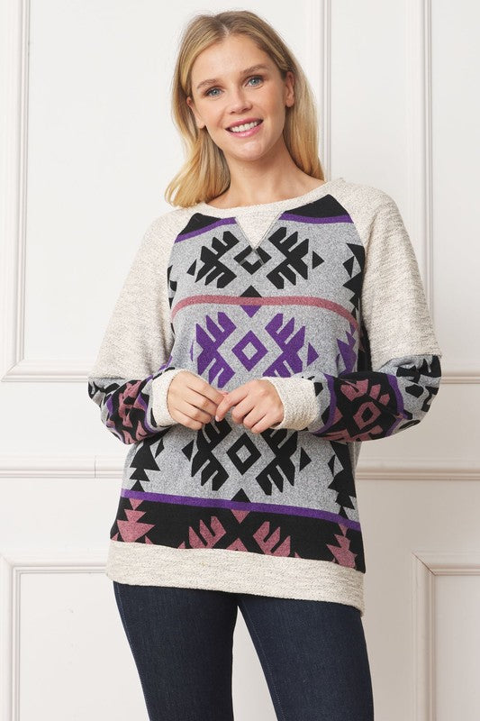 A smiling blonde-haired woman is wearing the V Stitch Tribal Contrast Tunic, which features geometric designs in purple, black, and pink, standing against a white background.