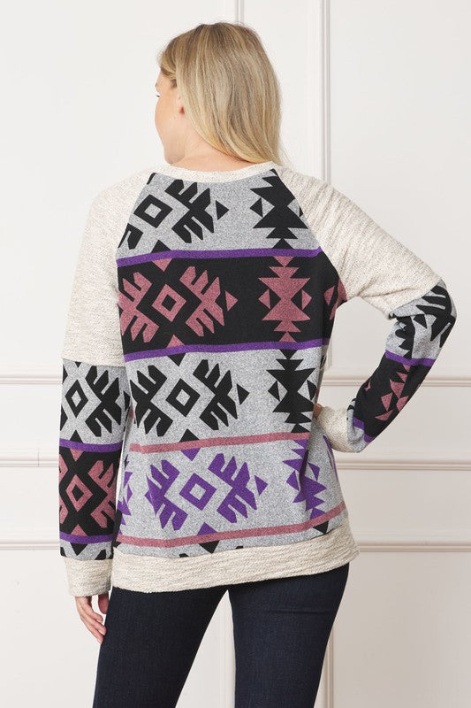 A woman stands smiling, wearing a Plus V Stitch Tribal Contrast Tunic with geometric patterns in black, grey, purple, and pink. The long-sleeve knit fabric is infused with gold thread. She has blonde hair and dark pants against a light-colored wall.