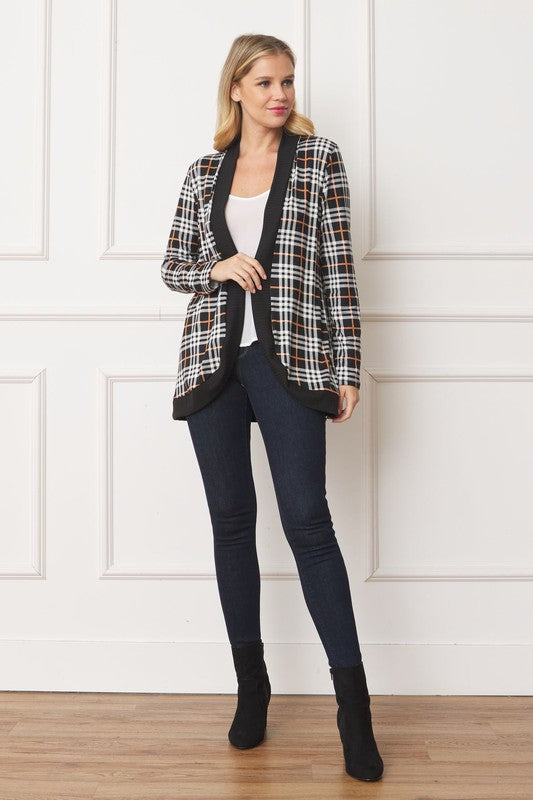 A woman with blonde hair stands against a white background, looking at the camera with a neutral expression. She is wearing the Plaid Contrast Drape Open Cardigan over a white top and dark jeans. Made in the United States, this cardigan features a stylish black and white plaid design.