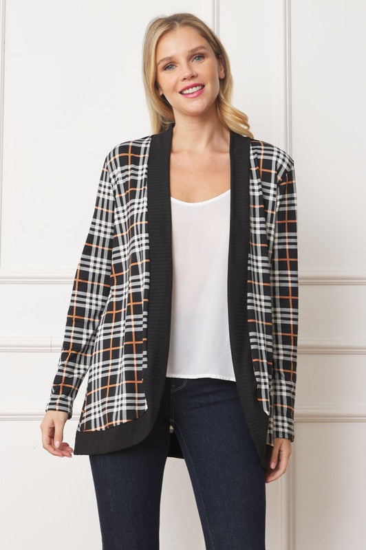 A woman with blonde hair stands against a white background, looking at the camera with a neutral expression. She is wearing the Plaid Contrast Drape Open Cardigan over a white top and dark jeans. Made in the United States, this cardigan features a stylish black and white plaid design.