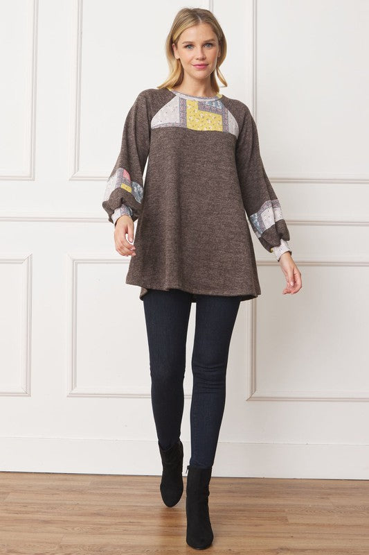A woman in a Patchwork Panel Accent A-Line Tunic, featuring a grey long-sleeve top with pink and blue patchwork panel details on the sleeves and chest, stands in front of a light-colored wall.