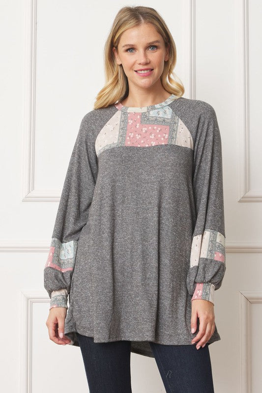 A woman in a Patchwork Panel Accent A-Line Tunic, featuring a grey long-sleeve top with pink and blue patchwork panel details on the sleeves and chest, stands in front of a light-colored wall.