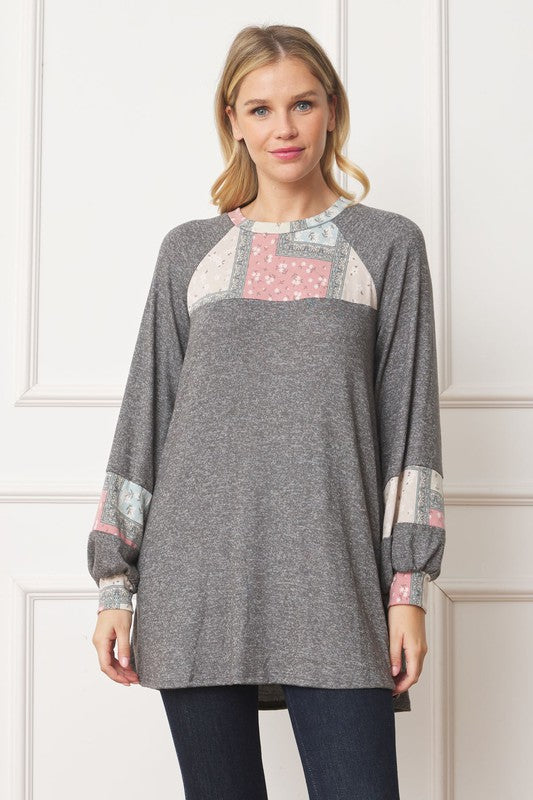 A woman in a Patchwork Panel Accent A-Line Tunic, featuring a grey long-sleeve top with pink and blue patchwork panel details on the sleeves and chest, stands in front of a light-colored wall.