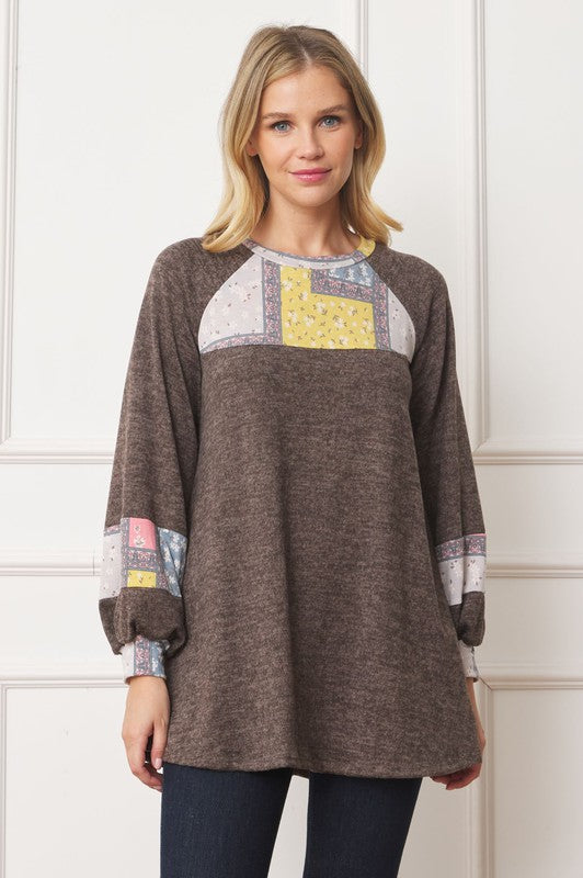 A woman in a Patchwork Panel Accent A-Line Tunic, featuring a grey long-sleeve top with pink and blue patchwork panel details on the sleeves and chest, stands in front of a light-colored wall.
