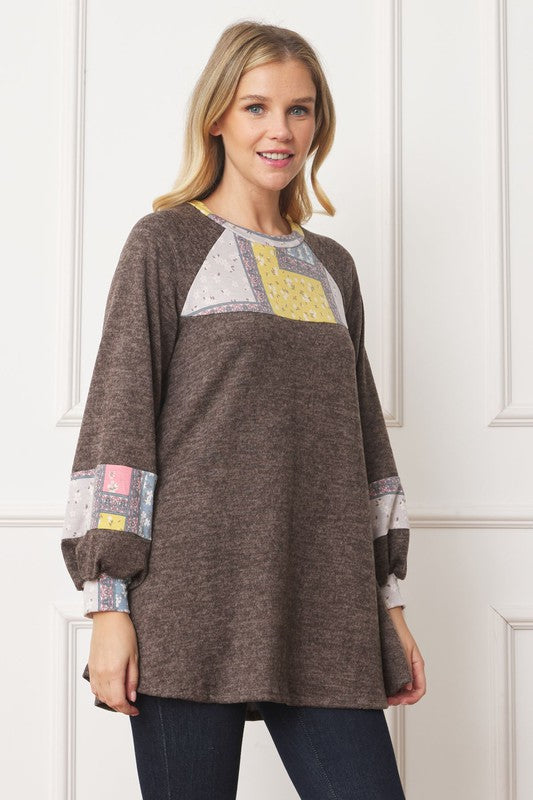 A woman in a Patchwork Panel Accent A-Line Tunic, featuring a grey long-sleeve top with pink and blue patchwork panel details on the sleeves and chest, stands in front of a light-colored wall.