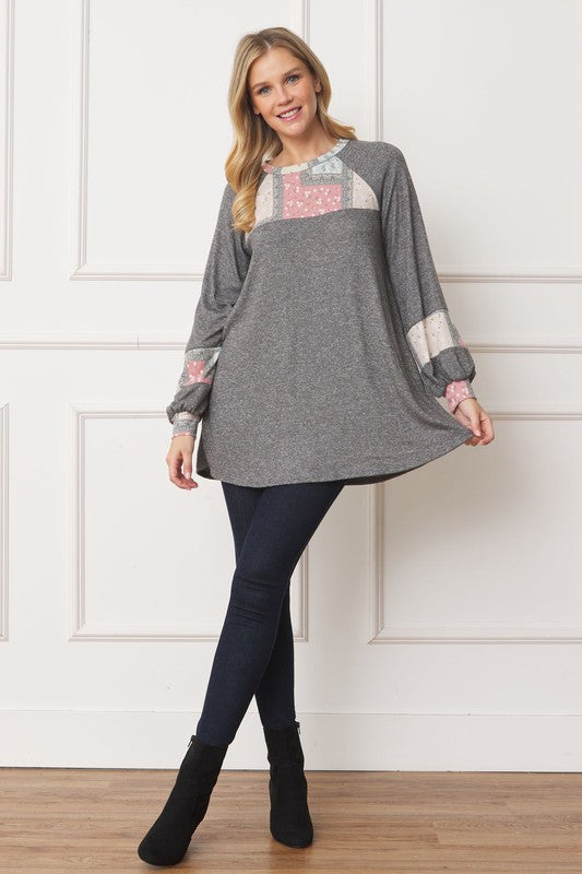 A woman in a Patchwork Panel Accent A-Line Tunic, featuring a grey long-sleeve top with pink and blue patchwork panel details on the sleeves and chest, stands in front of a light-colored wall.