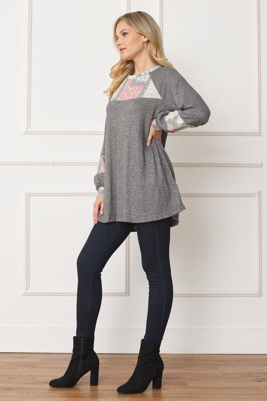 A woman in a Patchwork Panel Accent A-Line Tunic, featuring a grey long-sleeve top with pink and blue patchwork panel details on the sleeves and chest, stands in front of a light-colored wall.