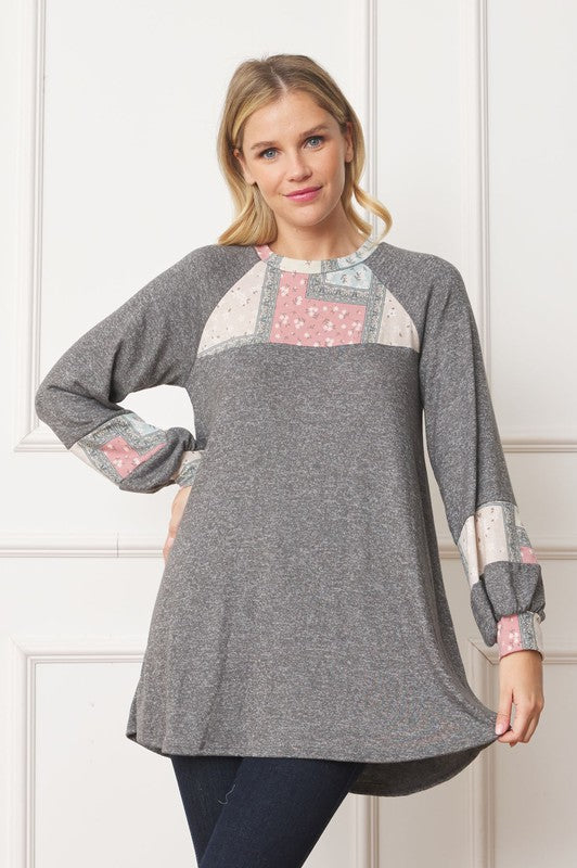 A woman in a Patchwork Panel Accent A-Line Tunic, featuring a grey long-sleeve top with pink and blue patchwork panel details on the sleeves and chest, stands in front of a light-colored wall.