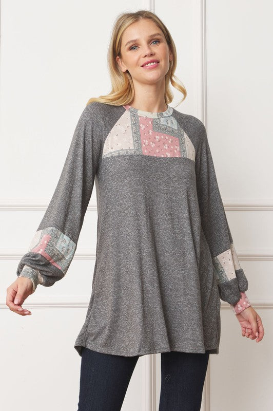 A woman in a Patchwork Panel Accent A-Line Tunic, featuring a grey long-sleeve top with pink and blue patchwork panel details on the sleeves and chest, stands in front of a light-colored wall.