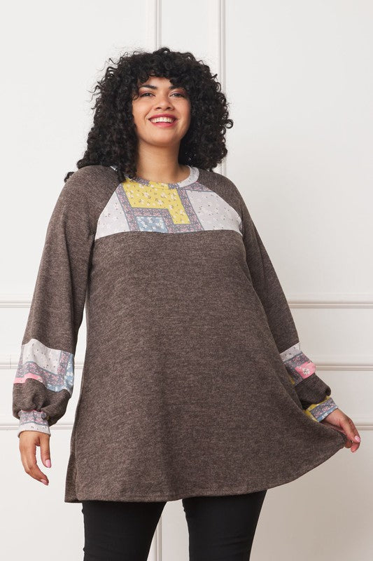 A woman with curly hair wears the Plus Patchwork Panel Accent A-Line Tunic in brown, featuring patterned details and patchwork panels on the chest and sleeves, standing against a white paneled background.