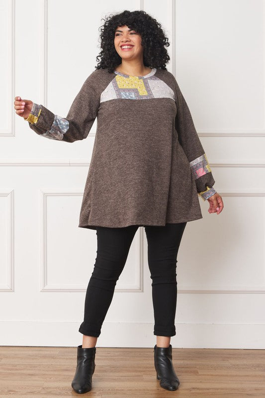 A person with curly hair is wearing a long-sleeved brown Plus Patchwork Panel Accent A-Line Tunic, standing in front of a white wall.