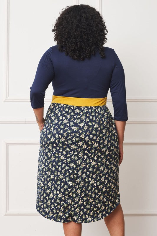 A woman with curly hair smiles, adorning a Plus Curved Hem Band Midi Dress made from a polyester and spandex blend. The black dress showcases three-quarter sleeves and is accented with a red waistband. The skirt is decorated with an eye-catching floral pattern of large pink and orange flowers.