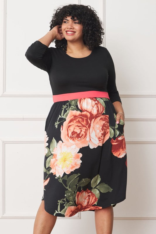 A woman with curly hair smiles, adorning a Plus Curved Hem Band Midi Dress made from a polyester and spandex blend. The black dress showcases three-quarter sleeves and is accented with a red waistband. The skirt is decorated with an eye-catching floral pattern of large pink and orange flowers.