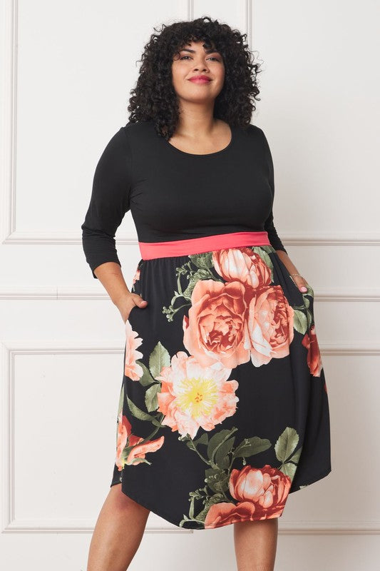 A woman with curly hair smiles, adorning a Plus Curved Hem Band Midi Dress made from a polyester and spandex blend. The black dress showcases three-quarter sleeves and is accented with a red waistband. The skirt is decorated with an eye-catching floral pattern of large pink and orange flowers.