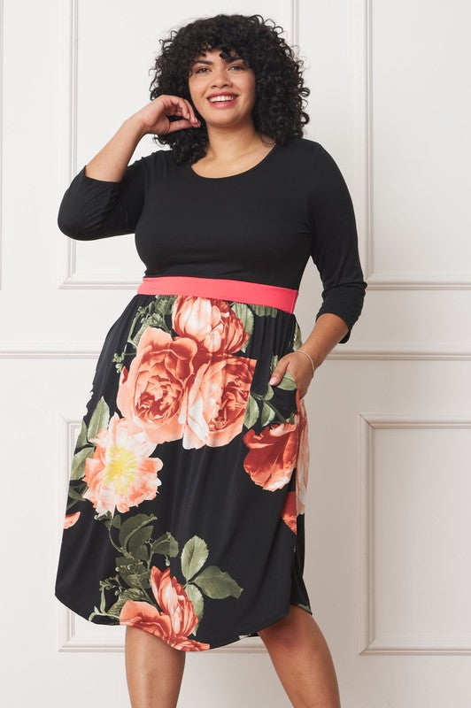 A woman with curly hair smiles, adorning a Plus Curved Hem Band Midi Dress made from a polyester and spandex blend. The black dress showcases three-quarter sleeves and is accented with a red waistband. The skirt is decorated with an eye-catching floral pattern of large pink and orange flowers.